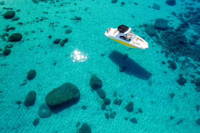 Aerial view of a white-and-yellow speedboat anchored in crystal-clear turquoise water, sun-speckled surface revealing scattered submerged rocks and the boat's dark shadow on the sandy seabed.