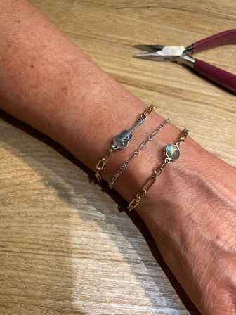 Close-up of a wrist wearing three mixed-metal bracelets: a gold link chain with an iridescent round stone, a delicate silver chain, and a gold chain with a tiny guitar charm, resting on a light wood tabletop with maroon-handled jewelry pliers nearby.