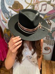 Bride-to-be in white dress holds a black wide-brim fedora with braided hatband, posed against a colorful music-themed mural in a boutique setting.