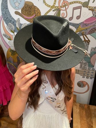 Bride-to-be in white dress holds a black wide-brim fedora with braided hatband, posed against a colorful music-themed mural in a boutique setting.