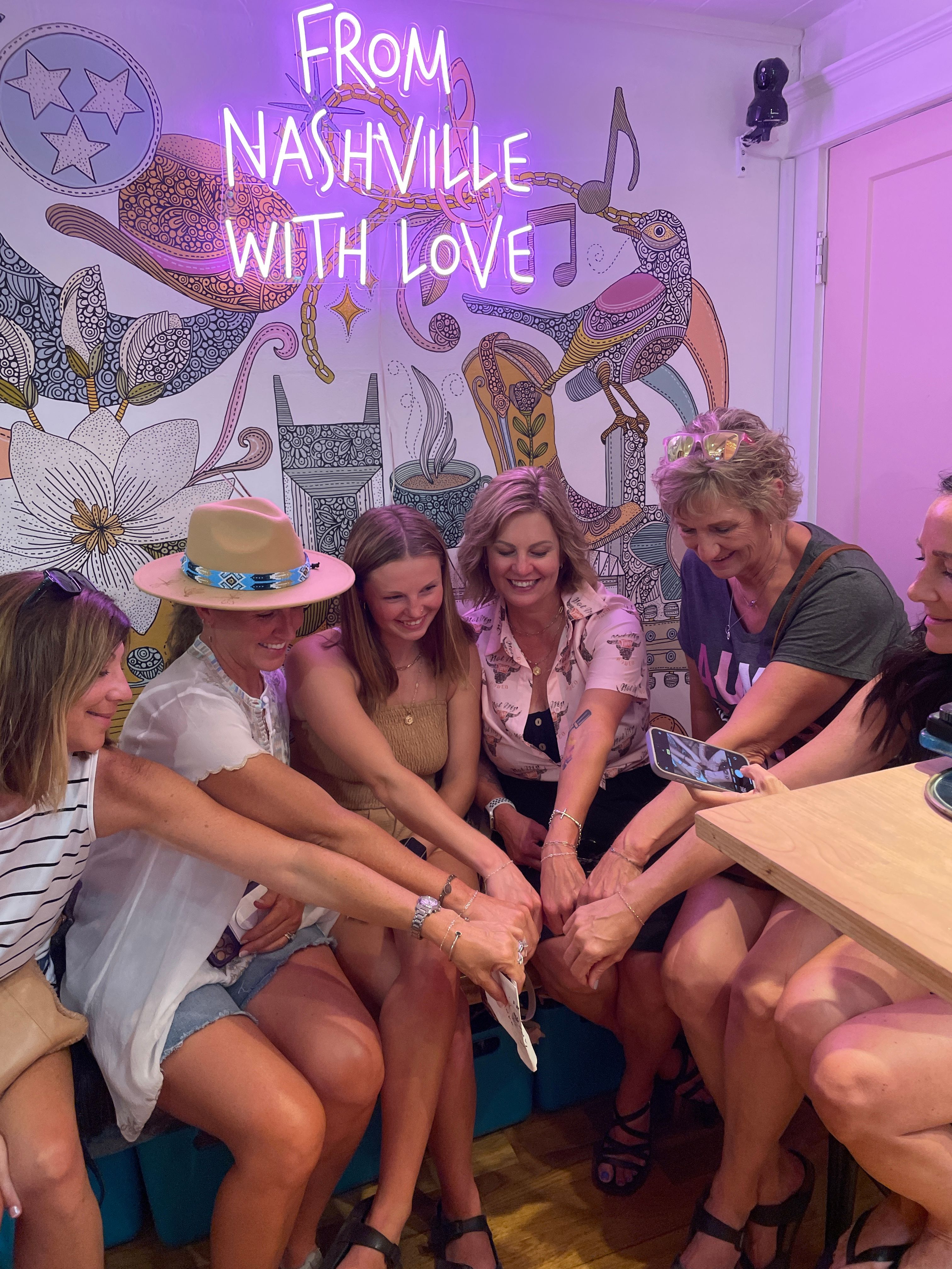 Smiling group of friends in a colorful Nashville shop leaning together to show matching bracelets, under a pink neon "From Nashville With Love" sign and decorative floral bird mural.