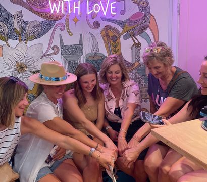 Smiling group of friends in a colorful Nashville shop leaning together to show matching bracelets, under a pink neon "From Nashville With Love" sign and decorative floral bird mural.