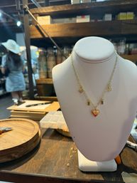 Dainty gold charm necklace with heart, sun and moon pendants displayed on a white bust on a wooden table at an artisan market jewelry booth, blurred shelves and shoppers in the background.