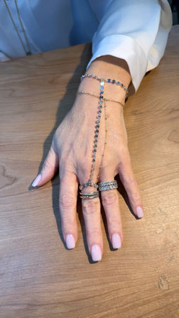 Close-up of a manicured hand with pale pink nails resting on a wooden table, wearing stacked diamond rings and delicate gold and silver bracelets connected by a sparkly hand chain.