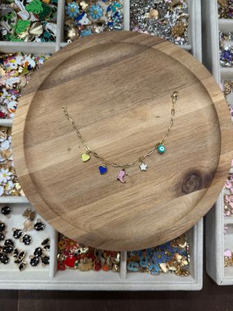 Playful gold charm bracelet on a round wooden tray featuring yellow and blue hearts, a pink boot, white star and turquoise eye charm, photographed on a jewelry display tray with assorted colorful enamel charms.