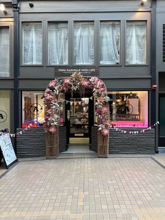 Charming boutique storefront in an indoor arcade with a wicker arch of pink and blush flowers and bunting over a glass door, flanked by display windows and a pink neon 'permanent jewelry' sign, tiled walkway in front.