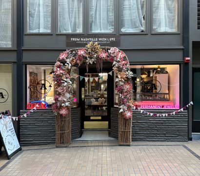 Charming boutique storefront in an indoor arcade with a wicker arch of pink and blush flowers and bunting over a glass door, flanked by display windows and a pink neon 'permanent jewelry' sign, tiled walkway in front.