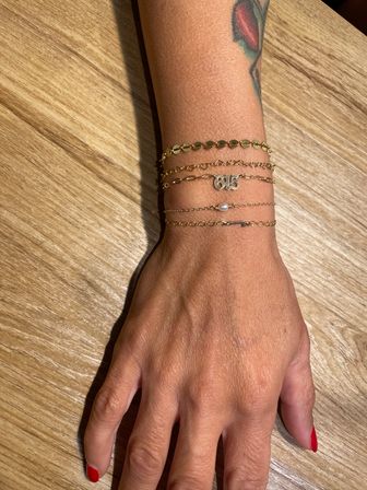 Close-up top-down view of a tattooed wrist and hand wearing stacked delicate gold chain bracelets — including a small pearl and a numeric charm — with red-painted nails on a light wood table.