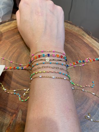 Wrist wearing a layered stack of delicate gold chains and tiny multicolored bead bracelets resting on a wooden surface — playful summer jewelry