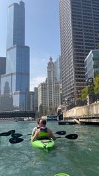 Two kayakers paddling on the Chicago River past downtown skyscrapers and a historic clock-topped tower under a clear blue sky