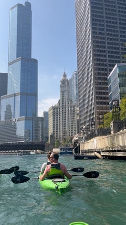 Two kayakers paddling on the Chicago River past downtown skyscrapers and a historic clock-topped tower under a clear blue sky