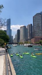 Kayakers in bright green kayaks paddling on the Chicago River near downtown, with tour boats and a skyline of high-rise buildings under a sunny blue sky