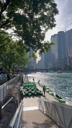 Green kayaks lined on a dock along the Chicago River with people boarding, a tree‑shaded riverwalk and downtown high‑rise skyline with sunlight reflecting on the water.
