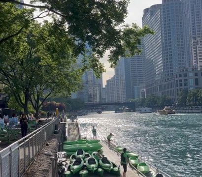 Green kayaks lined on a dock along the Chicago River with people boarding, a tree‑shaded riverwalk and downtown high‑rise skyline with sunlight reflecting on the water.