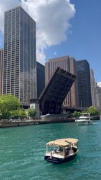 Small leisure boat on the turquoise Chicago River passing a raised drawbridge with downtown skyscrapers under a sunny blue sky