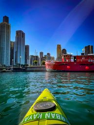 POV from a yellow kayak on teal water, approaching a red fireboat with Chicago skyscrapers and a vivid blue sky in the background.