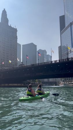 Two paddlers in a bright green kayak on the Chicago River, passing beneath a flag-lined downtown bridge with the sunlit Chicago skyline of glass skyscrapers.