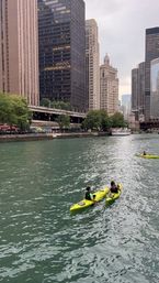 Two kayakers paddling bright yellow kayaks on the Chicago River past downtown Chicago skyscrapers and a riverboat under an overcast sky.