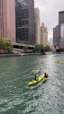 Two kayakers paddling bright yellow kayaks on the Chicago River past downtown Chicago skyscrapers and a riverboat under an overcast sky.