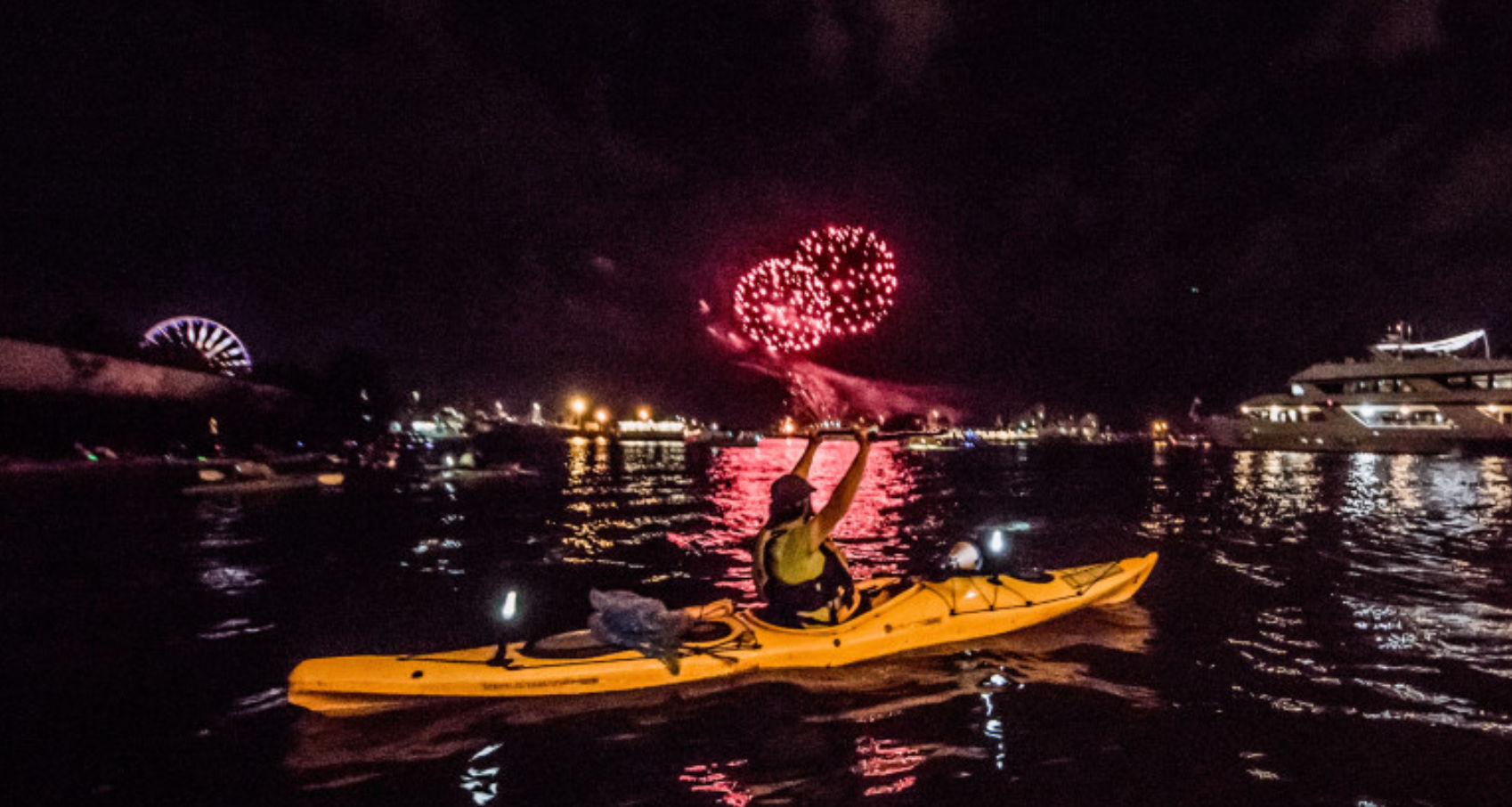 Kayaker in a yellow kayak at night watching red fireworks over a harbor, with a lit ferris wheel and yacht reflections on the water