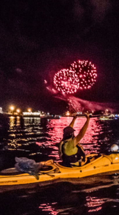 Kayaker in a yellow kayak at night watching red fireworks over a harbor, with a lit ferris wheel and yacht reflections on the water