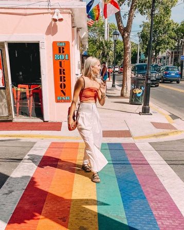 Woman in an orange crop top and white skirt walking across a rainbow-painted crosswalk on a sunny Key West street with pastel storefronts, palm trees, flags, and parked cars.