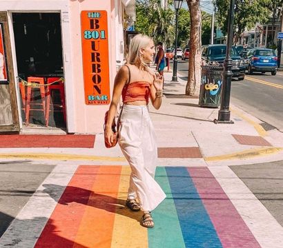 Woman in an orange crop top and white skirt walking across a rainbow-painted crosswalk on a sunny Key West street with pastel storefronts, palm trees, flags, and parked cars.