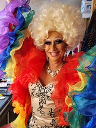 Smiling drag performer in a voluminous platinum wig and bold makeup, wearing a jeweled necklace and satin floral gown with vibrant rainbow ruffled sleeves.