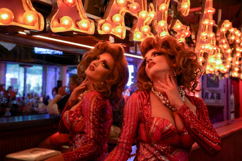 Glamorous drag performer in a red sequined dress and pearl necklace posing by a mirrored bar under warm marquee bulbs, dramatic makeup and reflection creating a neon-lit nightlife vibe.