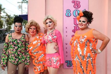 Four drag performers in vibrant sequined orange and pink costumes with dramatic makeup posing against a pastel-pink wall featuring 'Key West' lettering