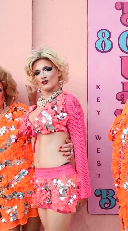 Four drag performers in vibrant sequined orange and pink costumes with dramatic makeup posing against a pastel-pink wall featuring 'Key West' lettering