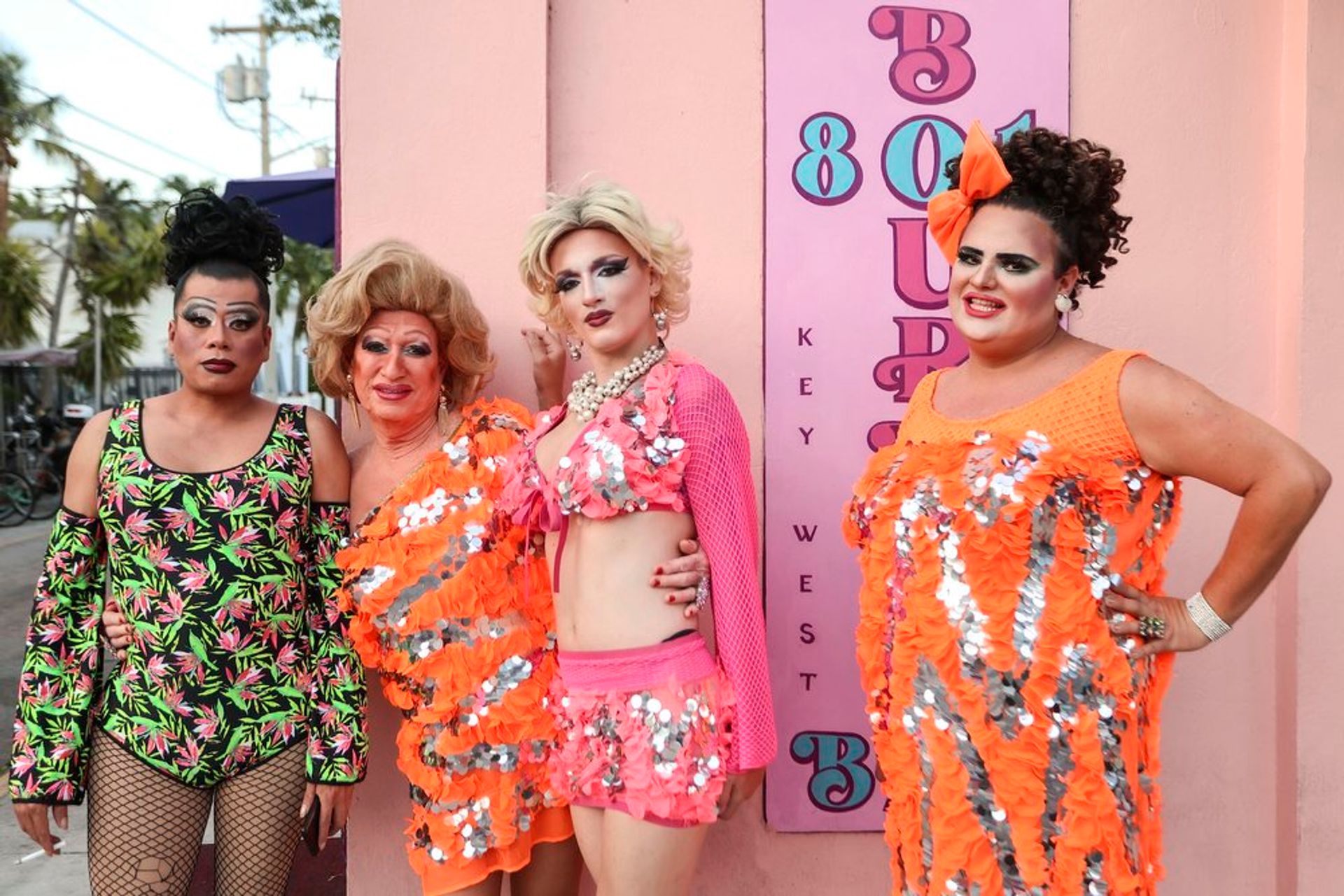 Four drag performers in vibrant sequined orange and pink costumes with dramatic makeup posing against a pastel-pink wall featuring 'Key West' lettering