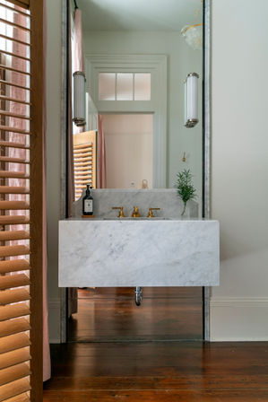 Chic marble floating bathroom sink with brass faucets and small plant, large mirror reflecting a doorway, wooden shutters and warm hardwood floor