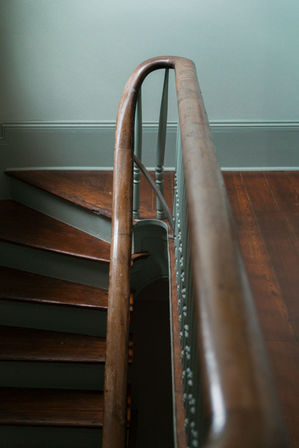 Close-up of a vintage wooden staircase interior featuring a curved polished banister, painted balusters, and worn hardwood treads