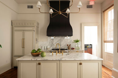 Sunlit luxury kitchen with marble island, brass faucet and chandelier, cream cabinetry, black curved range hood and a wooden bowl of watermelons.
