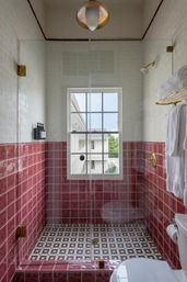 Glass-enclosed walk-in shower with glossy rose-pink lower subway tiles and white upper tiles, mosaic tiled floor, brass fixtures, wall soap dispensers, a window view and white towels on a gold rack.