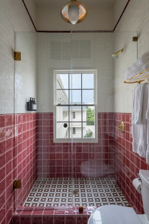 Glass-enclosed walk-in shower with glossy rose-pink lower subway tiles and white upper tiles, mosaic tiled floor, brass fixtures, wall soap dispensers, a window view and white towels on a gold rack.