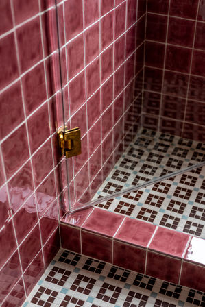 Retro pink ceramic-tiled shower corner with white-grouted walls, brown-and-blue mosaic floor and a glass door with brass hinge