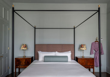 Four-poster canopy bed with patterned headboard, crisp white linens and blue bolster, wooden nightstands with floral lamps and a striped robe against pale gray walls.
