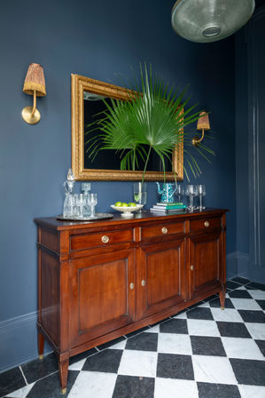 Navy entryway with a vintage wooden sideboard on black-and-white checkered marble floor, ornate gold-framed mirror, large palm fan in a glass vase, crystal decanter and glasses, stacked books and brass wall sconces.