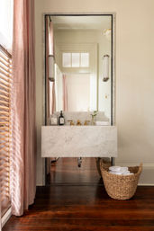 Chic bathroom with a floating marble sink and brass faucets, tall mirror reflecting a doorway, soft pink linen curtains and wooden shutters, dark hardwood floor and woven basket of white towels.