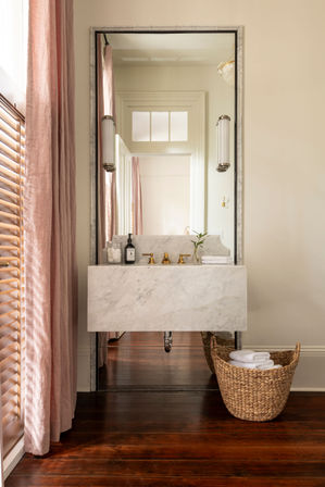 Chic bathroom with a floating marble sink and brass faucets, tall mirror reflecting a doorway, soft pink linen curtains and wooden shutters, dark hardwood floor and woven basket of white towels.