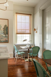 Sunlit kitchen breakfast nook with striped banquette, green French-style rattan bistro chairs, small round wooden table, bamboo roman shade, marble countertop edge and warm hardwood floors.