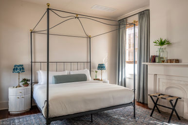 Cozy sunlit bedroom interior with black metal four‑poster canopy bed, crisp white bedding and teal bolster pillow, blue patterned bedside lamps, gray curtains, decorative mantel with vase and books, and a patterned rug on hardwood floors.