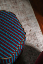 Close-up of an upholstered ottoman with bold blue and maroon diagonal stripes and contrasting piped trim, placed on a faded area rug beside a hardwood floor