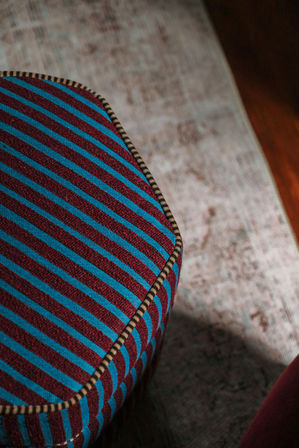 Close-up of an upholstered ottoman with bold blue and maroon diagonal stripes and contrasting piped trim, placed on a faded area rug beside a hardwood floor