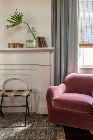 Cozy living room corner with a pink velvet armchair next to a white decorative mantel topped by a glass jar with a large green leaf, a small stack of books, grey curtains, bamboo blind, woven stool and patterned rug.