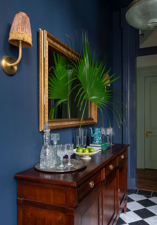 Navy-painted foyer with a mahogany sideboard under a gilded mirror, tall palm fronds in a vase, crystal decanter and glasses, a bowl of limes, vintage fringed wall sconce and black-and-white checkered floor — stylish home entry vignette.