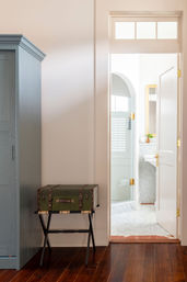 Bright home entry with a green vintage trunk on a folding luggage rack beside a blue‑gray wardrobe, dark hardwood floor and an open white door revealing a marble bathroom with glass shower, arched window and gold hardware.