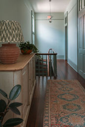 Cozy upstairs hallway with dark hardwood floors, muted green walls, an antique sideboard topped with a terracotta lamp and potted plant, patterned runner rug, and a curved wooden banister leading to stairs.